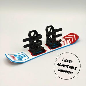 May include: A miniature white snowboard with red and blue accents, featuring black adjustable bindings. The board has the text "LIL BRO USA" and a red arrow design. A circular graphic states "I HAVE ADJUSTABLE BINDINGS!"