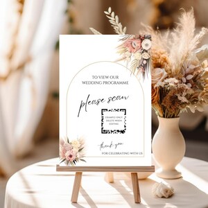 Könnte beinhalten: Ein weißes Hochzeitsschild auf einer kleinen Holzstaffelei mit dem Text „To View Our Wedding Programme, please scan“. Das Schild enthält einen QR-Code und florale Akzente in Rosa und Beige.