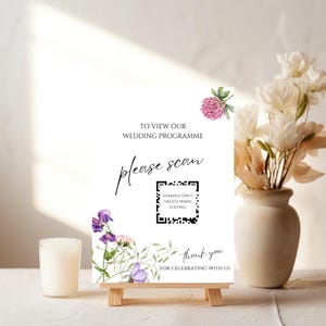 May include: A white wedding program sign on a small wooden easel. The sign has floral accents and text that reads "TO VIEW OUR WEDDING PROGRAMME, please scan" with a QR code. The sign also says "thank you for celebrating with us."