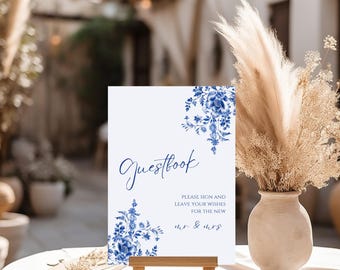 May include: A white sign with blue floral accents and the word "Guestbook" in elegant script. The sign also includes the text "Please sign and leave your wishes for the new mr. & mrs." It is displayed on a small wooden easel.