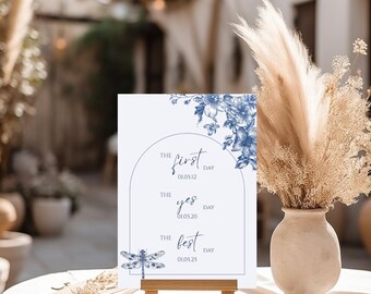 May include: A white sign on a wooden easel with blue floral accents and text. The sign reads "The First Day," "The Yes Day," and "The Best Day," with corresponding dates. A dragonfly illustration is at the bottom.