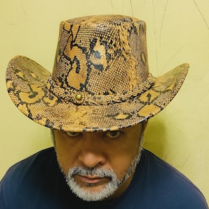 Może przedstawiać: Kapelusz kowbojski z wzorem skóry węża w odcieniach brązu i czerni. Kapelusz ma szerokie rondo i wysoką koronę, z plecionym paskiem wokół podstawy korony. Z przodu kapelusza znajduje się mosiężny guzik.