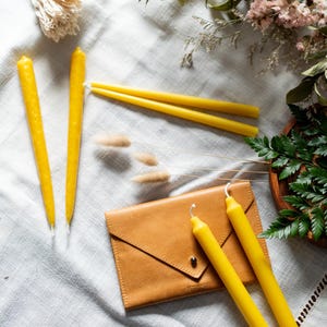 Puede incluir: Un conjunto de velas cónicas amarillas, un sobre de cuero marrón y vegetación dispuesta sobre una superficie de lino blanco. Las velas son de diferentes longitudes y no están encendidas. También hay flores secas y un plato de madera.