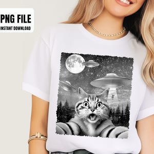 May include: White t-shirt featuring a black and white graphic of a surprised cat taking a selfie with UFOs and a full moon in the background. The text "PNG FILE INSTANT DOWNLOAD" is in the upper left corner.