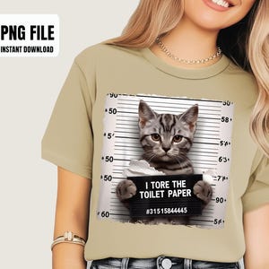 May include: A beige t-shirt with a humorous design. It features a grey tabby cat in a mugshot pose, holding a sign saying "I TORE THE TOILET PAPER." The shirt also has the text "PNG FILE INSTANT DOWNLOAD".
