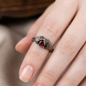 Può includere: Un anello in argento scuro con una pietra preziosa rossa. Il design dell'anello include due mani stilizzate che cullano la gemma. L'anello è indossato su un dito, mettendo in risalto i dettagli intricati e il colore vibrante della pietra.