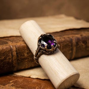 Puede incluir: Un anillo de estilo antiguo con una gran piedra preciosa ovalada de color púrpura oscuro engastada en una banda de metal ornamentada de color bronce. El anillo se exhibe sobre un palillo de madera de color claro, sobre un fondo de libros y pergamino antiguos.