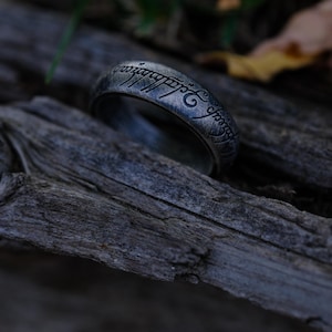 Può includere: Un anello d'argento con scritta incisa, appoggiato su legno stagionato. La superficie dell'anello è dettagliata con scritte intricate e ha un aspetto leggermente usurato. È un gioiello.