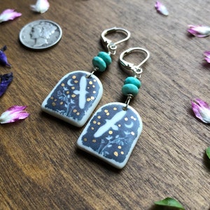May include: A pair of silver dangle earrings with turquoise beads and a ceramic charm featuring a white bird flying against a dark blue background with gold accents.