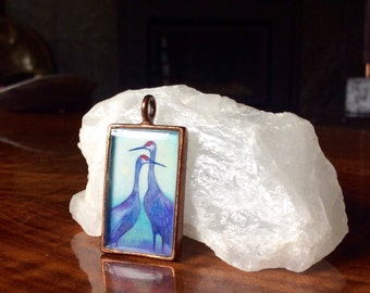 May include: A rectangular pendant with a copper-colored frame features a painting of two blue birds with red accents. The pendant is displayed on a reflective wooden surface, next to a large, white, crystalline rock. The pendant has a small loop at the top.