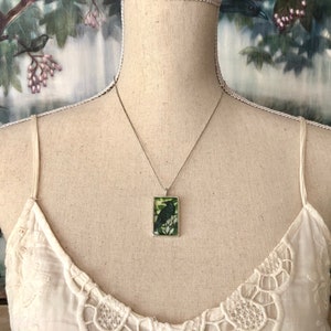 Puede incluir: Collar plateado con un colgante rectangular con una ilustraci&oacute;n de un p&aacute;jaro negro sobre un fondo verde. El colgante cuelga de una cadena fina. El collar se muestra en un maniqu&iacute; que lleva una camiseta sin mangas blanca.