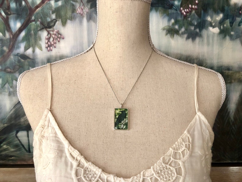 Puede incluir: Collar plateado con un colgante rectangular con una ilustraci&oacute;n de un p&aacute;jaro negro sobre un fondo verde. El colgante cuelga de una cadena fina. El collar se muestra en un maniqu&iacute; que lleva una camiseta sin mangas blanca.