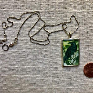 Puede incluir: Collar plateado con un colgante rectangular que presenta un cuervo verde oscuro sobre flores blancas sobre un fondo verde y amarillo. Una moneda est&aacute; al lado del colgante para la comparaci&oacute;n de tama&ntilde;o.