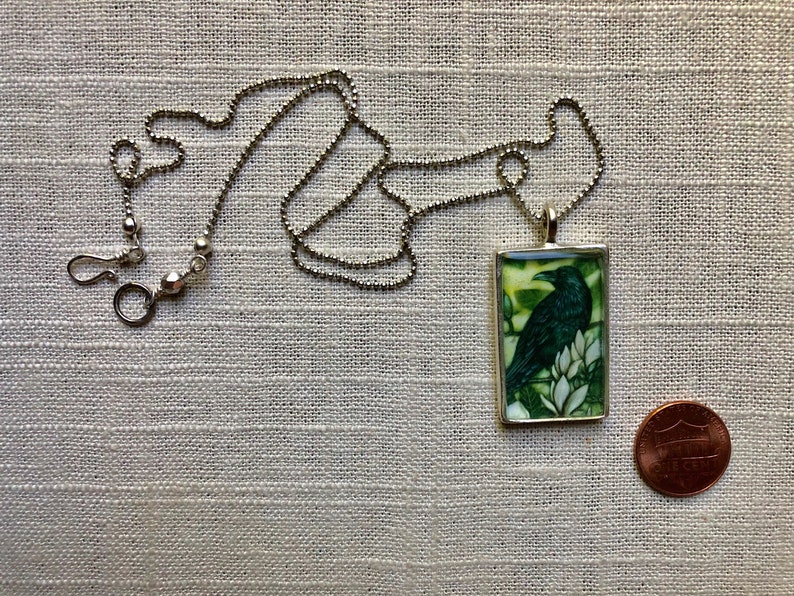 Puede incluir: Collar plateado con un colgante rectangular que presenta un cuervo verde oscuro sobre flores blancas sobre un fondo verde y amarillo. Una moneda est&aacute; al lado del colgante para la comparaci&oacute;n de tama&ntilde;o.