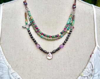 Puede incluir: Collar de varias hebras con cuentas verdes, rosas y negras. Incluye un colgante plateado y un dije con diseño de águila. El collar se exhibe sobre una superficie blanca. La longitud del collar es de unos 40 cm.