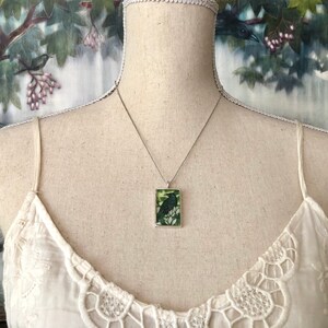 Puede incluir: Collar plateado con un colgante rectangular con un dise&ntilde;o de p&aacute;jaro negro sobre un fondo verde y blanco. El colgante cuelga de una delicada cadena. El collar se exhibe en un maniqu&iacute; con un top de encaje blanco.