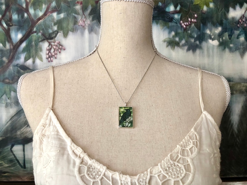 Puede incluir: Collar plateado con un colgante rectangular con un dise&ntilde;o de p&aacute;jaro negro sobre un fondo verde y blanco. El colgante cuelga de una delicada cadena. El collar se exhibe en un maniqu&iacute; con un top de encaje blanco.