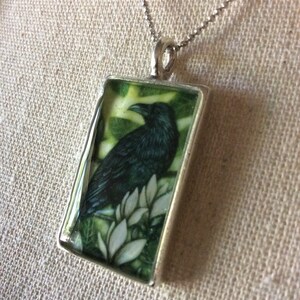 Puede incluir: Collar con colgante de plata. El colgante rectangular presenta una ilustraci&oacute;n detallada de un cuervo negro posado sobre flores blancas sobre un fondo verde. El colgante cuelga de una delicada cadena de plata.