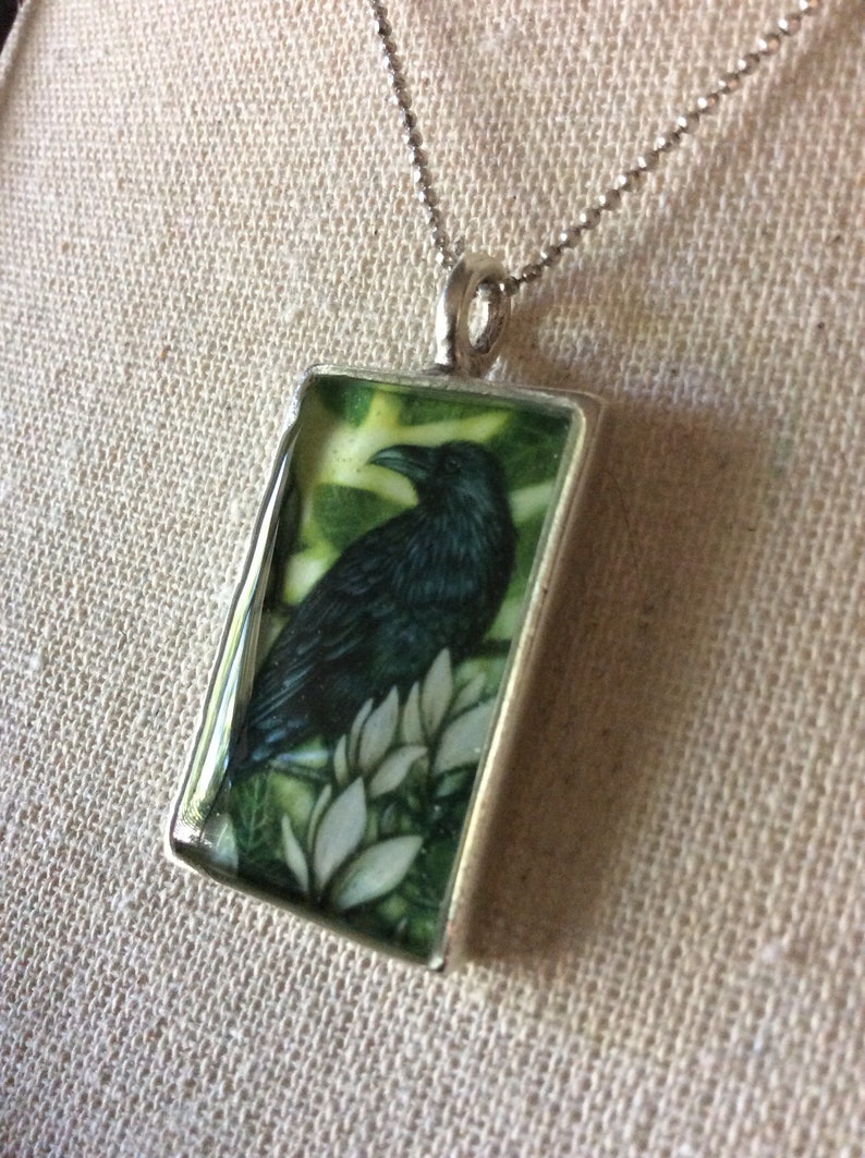 Puede incluir: Collar con colgante de plata. El colgante rectangular presenta una ilustraci&oacute;n detallada de un cuervo negro posado sobre flores blancas sobre un fondo verde. El colgante cuelga de una delicada cadena de plata.
