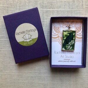 Puede incluir: Colgante rectangular con una ilustraci&oacute;n de un cuervo oscuro, en un marco plateado. El colgante se muestra en una caja de regalo morada con el texto "Rachelle Montoya Art Studio". La caja tambi&eacute;n tiene un logotipo de flor de loto.