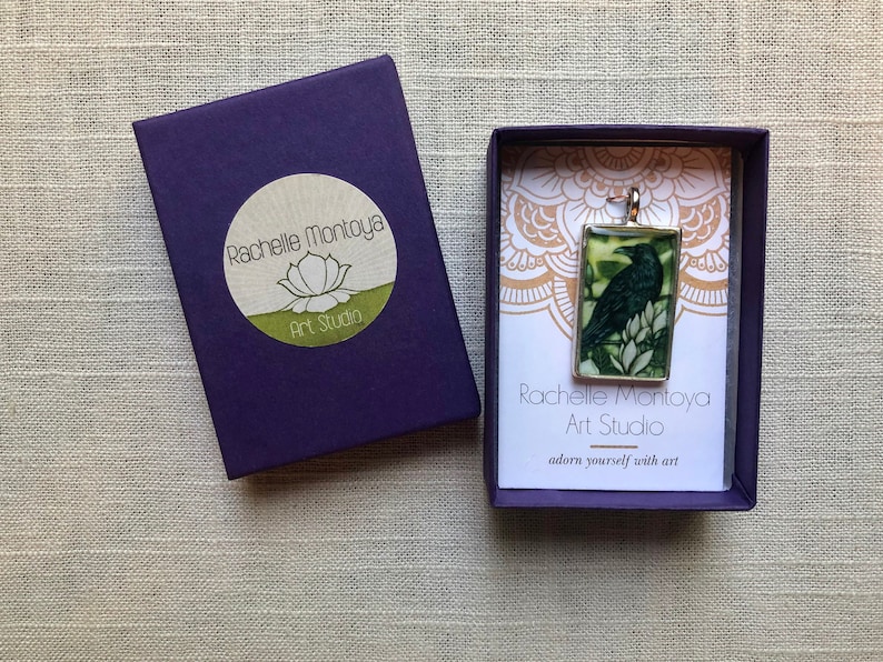 Puede incluir: Colgante rectangular con una ilustraci&oacute;n de un cuervo oscuro, en un marco plateado. El colgante se muestra en una caja de regalo morada con el texto "Rachelle Montoya Art Studio". La caja tambi&eacute;n tiene un logotipo de flor de loto.
