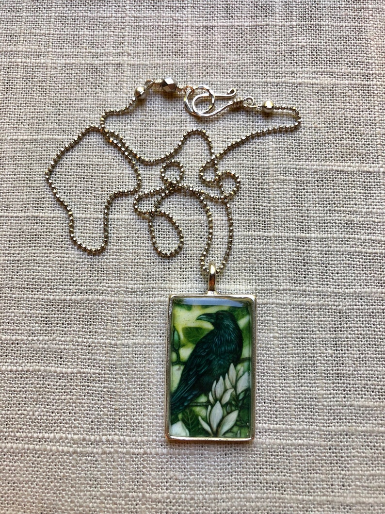 Puede incluir: Collar dorado con colgante rectangular. El colgante muestra un cuervo verde oscuro sobre flores blancas sobre un fondo verde y amarillo. La cadena es de estilo de cuentas.