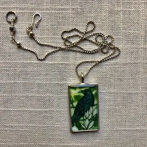 Puede incluir: Collar plateado con un colgante rectangular con una ilustraci&oacute;n de un cuervo verde oscuro. El cuervo est&aacute; posado sobre flores blancas sobre un fondo verde y amarillo. La cadena tiene un dise&ntilde;o de bolas y un cierre de mosquet&oacute;n.