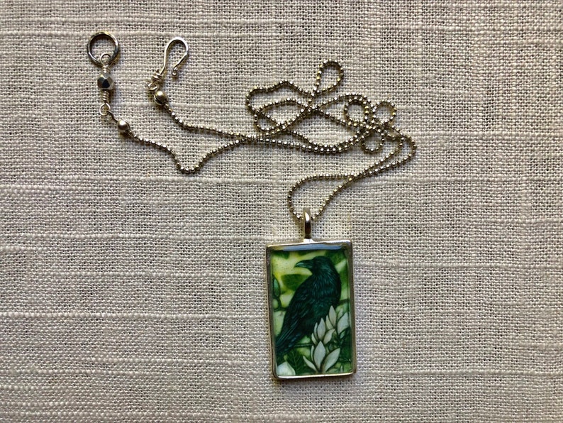 Puede incluir: Collar plateado con un colgante rectangular con una ilustraci&oacute;n de un cuervo verde oscuro. El cuervo est&aacute; posado sobre flores blancas sobre un fondo verde y amarillo. La cadena tiene un dise&ntilde;o de bolas y un cierre de mosquet&oacute;n.