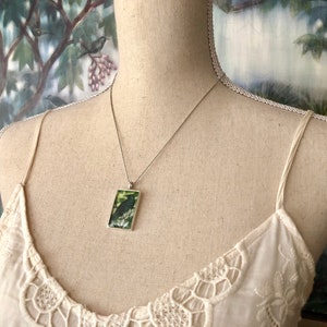 Puede incluir: Collar plateado con un colgante rectangular que presenta un p&aacute;jaro negro posado en una rama sobre un fondo verde y blanco. El colgante cuelga de una cadena fina, exhibido en un maniqu&iacute;.