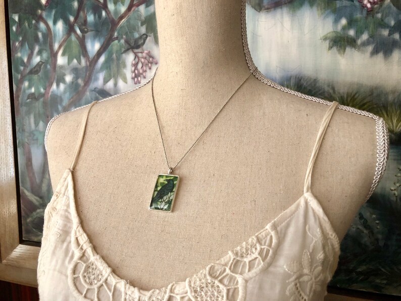 Puede incluir: Collar plateado con un colgante rectangular que presenta un p&aacute;jaro negro posado en una rama sobre un fondo verde y blanco. El colgante cuelga de una cadena fina, exhibido en un maniqu&iacute;.