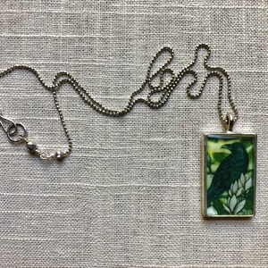 Puede incluir: Collar plateado con un colgante rectangular que presenta un cuervo negro posado sobre flores blancas sobre un fondo verde. El collar tiene una cadena de bolas y un cierre de mosquet&oacute;n.