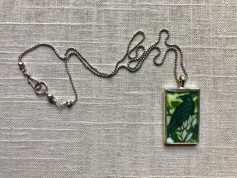 Puede incluir: Collar plateado con un colgante rectangular que presenta un cuervo negro posado sobre flores blancas sobre un fondo verde. El collar tiene una cadena de bolas y un cierre de mosquet&oacute;n.