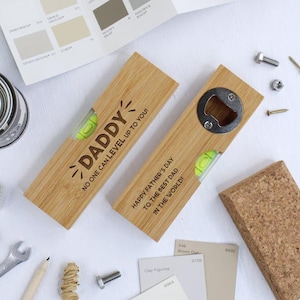 May include: Two wooden bottle openers with a spirit level and the text "DADDY" and "HAPPY FATHER'S DAY". One has a bottle opener. The openers are light brown with a yellow level bubble. Paint swatches, tools, and a cork are in the background.