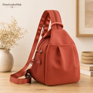 May include: A terracotta-coloured sling backpack featuring a single adjustable shoulder strap, multiple zippered compartments with circular pull tabs, and a smooth, solid-coloured exterior. The bag has a structured, rounded shape with a front pocket.