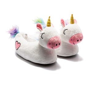 Puede incluir: Un par de zapatillas de unicornio de felpa blanca. Cada zapatilla presenta una nariz rosa, ojos cerrados, un cuerno dorado y una melena y cola coloridas. Una zapatilla tiene un corazón rosa en el lateral.