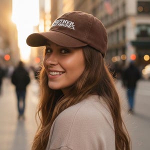 May include: A brown baseball cap with the text "TOGETHER AMERICA" in white. The cap has a curved brim and is worn by a person. The background is a city street with blurred buildings and people.