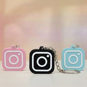 May include: Three Instagram-themed keychains in pink, black, and blue. Each keychain features a square design with a white outline and a white circle in the center, attached to a silver chain and keyring. The background is a soft gradient of pink and peach.