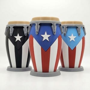 May include: Three conga drums featuring a flag-inspired design with a white star on a triangular field and vertical stripes. The drums have a tan top rim, grey hardware, and a grey circular base. The colors include black, white, red, and light blue.