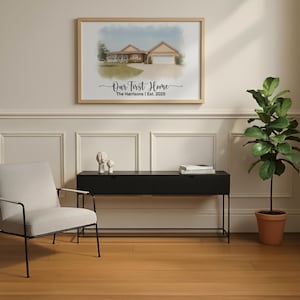 May include: A framed watercolor print of a house hangs on a wall. The print is in a light wood frame and features the text "Our First Home" and the family name and year. A black console table and a potted plant are below the print.