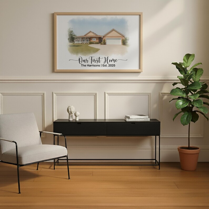 May include: A framed watercolor print of a house hangs on a wall. The print is in a light wood frame and features the text "Our First Home" and the family name and year. A black console table and a potted plant are below the print.