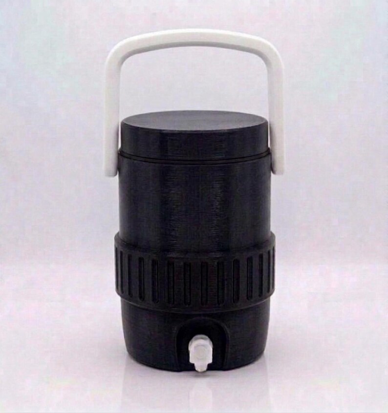 May include: A cylindrical black beverage cooler featuring a solid white arched handle attached to the top. The container has a textured ribbed band around the center and a small white spigot located at the bottom front for dispensing liquids.