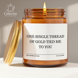 May include: A lit, amber-colored candle in a glass jar with a gold lid. The label reads "ONE SINGLE THREAD OF GOLD TIED ME TO YOU." The candle is 9 oz and made with 100% natural soy wax. The brand name is Célestia.