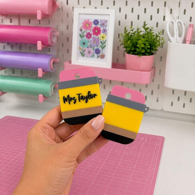 May include: Two pencil-shaped acrylic keychains featuring a pink eraser top, a grey metal band, a yellow body, and a black tip. One keychain displays the black script text Mrs Taylor on the yellow section. Both items have a small circular hole.