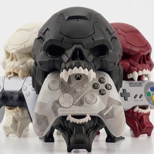 May include: A collection of skull-shaped gaming controller holders in white, black, and red. The holders are designed to resemble skulls with open mouths, holding various game controllers, including an Xbox controller with "PROJECT SCARLETT" text.
