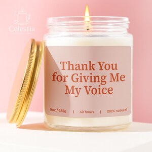 May include: A lit candle in a clear glass jar with a gold lid. The label reads "Thank You for Giving Me My Voice." The candle is 9oz and burns for 40 hours. The brand name "Célestia" is at the top left.