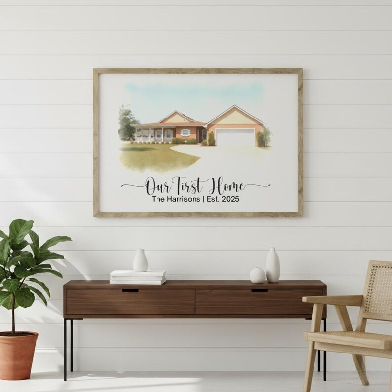 May include: Framed watercolor print of a house with a garage, set against a blue sky. The text "Our First Home" is written in cursive, with "The Harrisons | Est. 2025" below. The frame is a light brown color.