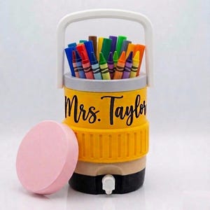 May include: A yellow cylindrical container with a white handle, a black base, and a spigot, filled with multicolored crayons. The text Mrs. Taylor is printed in black script on the side. A separate, round, light pink lid sits to the left.