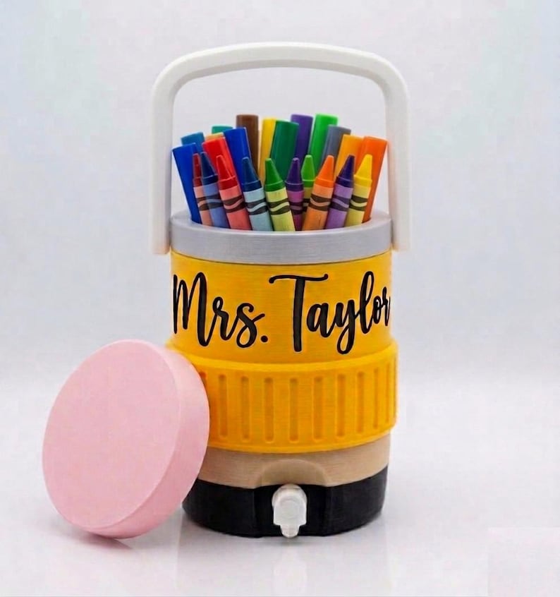 May include: A yellow cylindrical container with a white handle, a black base, and a spigot, filled with multicolored crayons. The text Mrs. Taylor is printed in black script on the side. A separate, round, light pink lid sits to the left.
