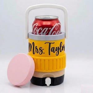 May include: A yellow and black beverage cooler with a white handle and a spigot, featuring the text Mrs. Taylor in black script. A red Coca-Cola can sits inside the top opening, and a separate round light pink lid is placed to the left side.