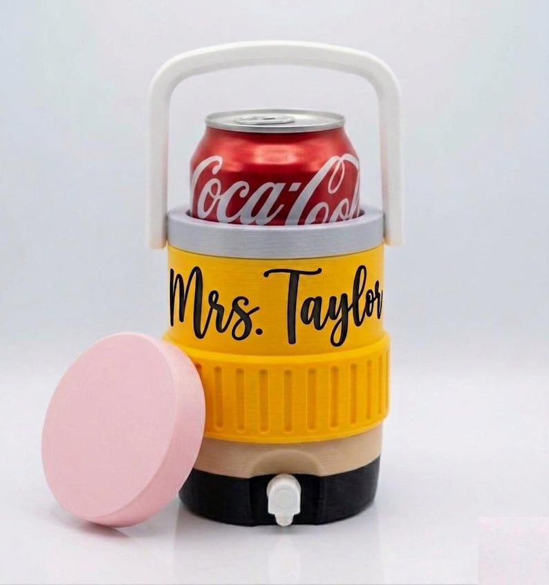 May include: A yellow and black beverage cooler with a white handle and a spigot, featuring the text Mrs. Taylor in black script. A red Coca-Cola can sits inside the top opening, and a separate round light pink lid is placed to the left side.